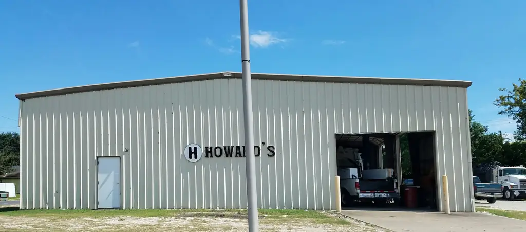 Cream corrugated steel shop building with a round H sign and the HOWARD'S name on the wall, an open roll-up bay with a white work truck inside, and heavy trucks parked at the right